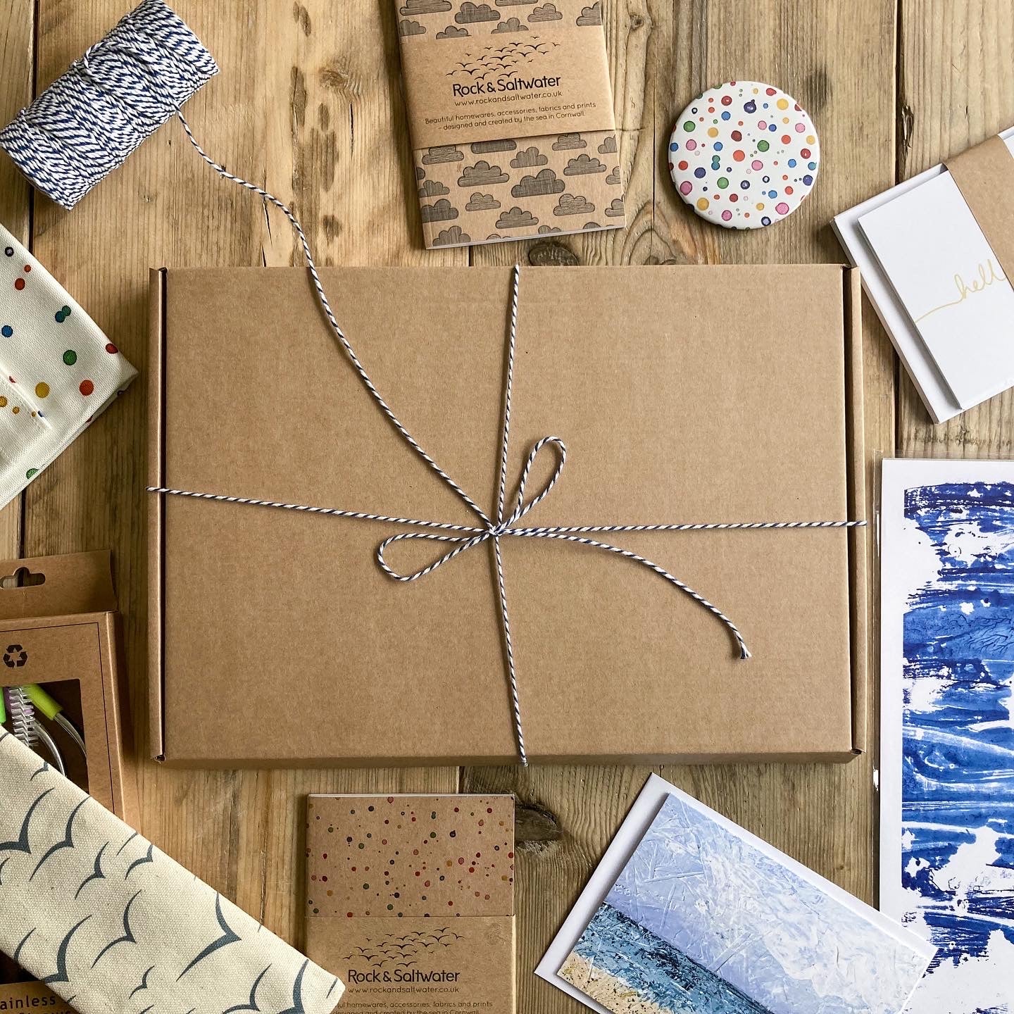 Brown cardboard gift box tied with navy blue baker's twine on a rustic wood table. Around the box are various Rock and Saltwater products including an cloud print notebook, paint print mirror, set of notecards, seascape print, greetings card and fabric.