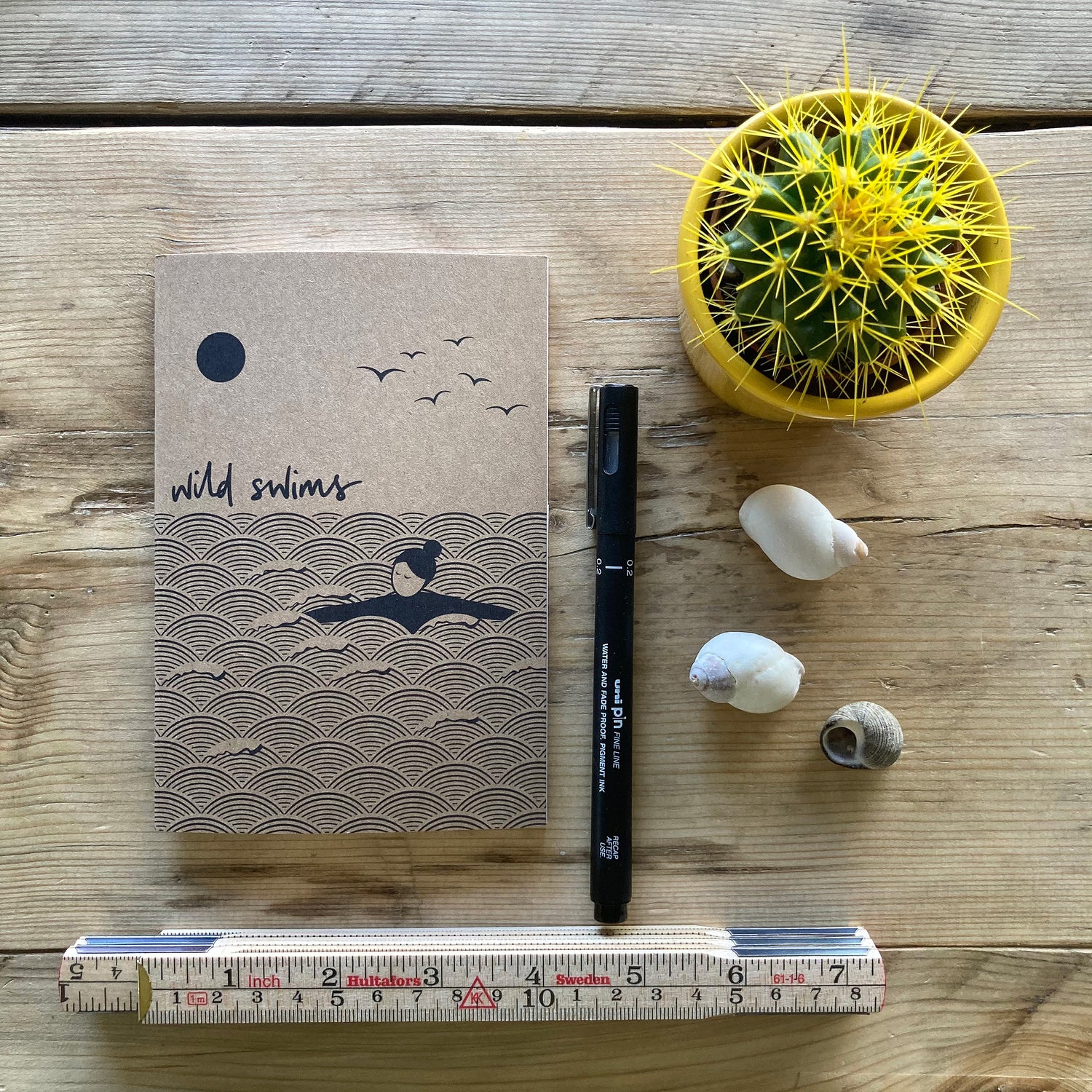 An A6 size wild swimming notebook sits on a rustic wooden table surrounded by a pen, wooden rule, three shells and a cacti in a yellow ceramic pot.