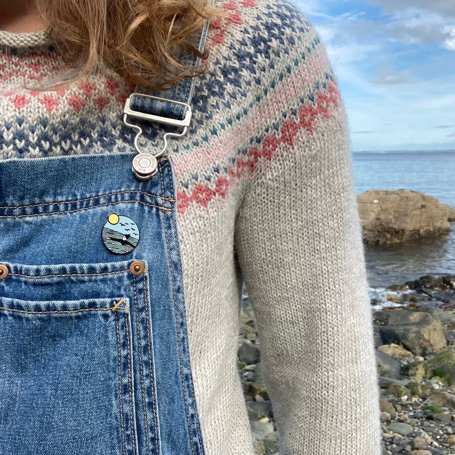 Close up of wild swimming enamel pin badge, attached to a pair of denim dungarees with a handknit fairisle jumper underneath. The sea can be seen behind the figure.