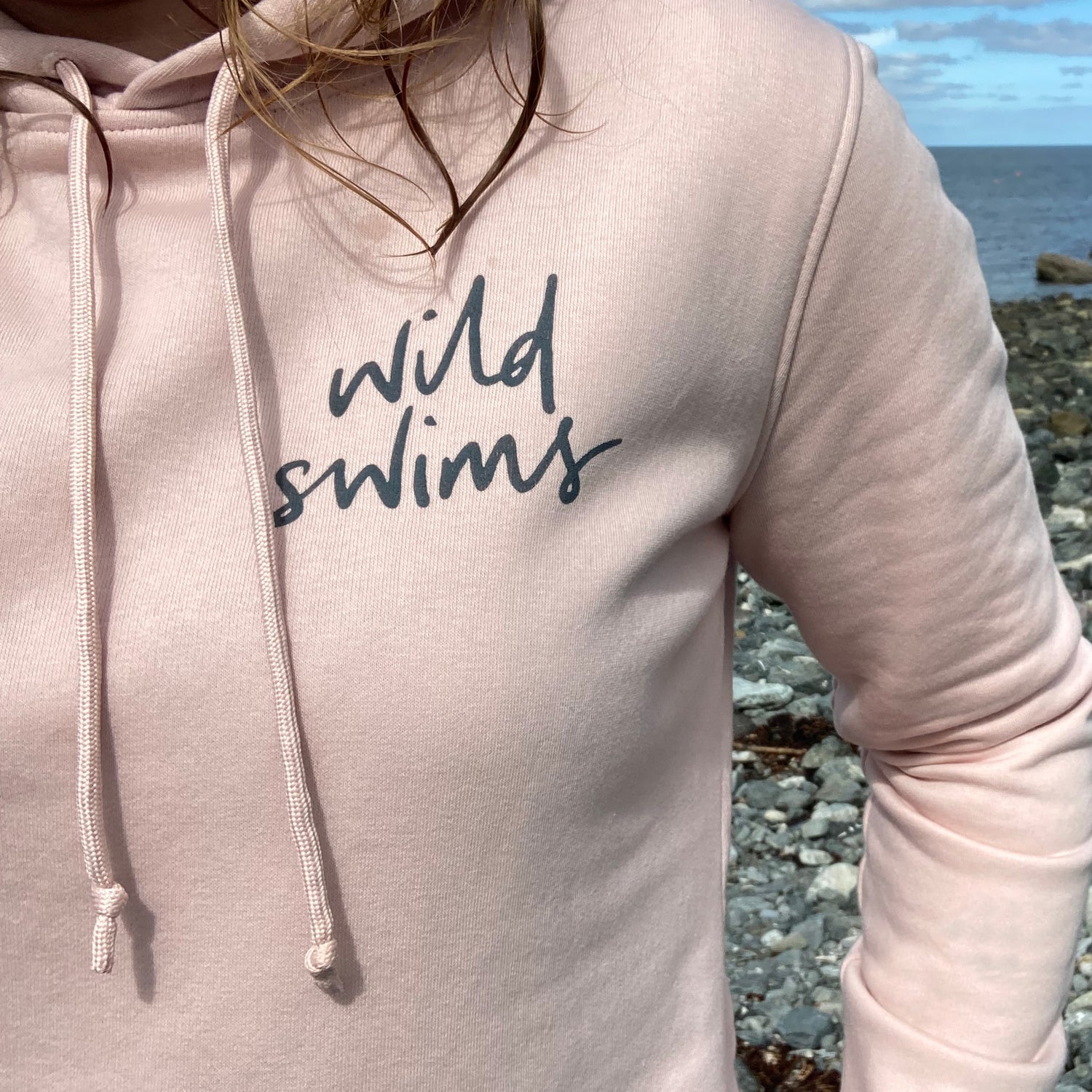 Cropped image of a women wearing a pink 'wild swims' hoodie. Her face is cropped out and the front print of the sweatshirt is visible. Behind her is a pebble beach and the sea.
