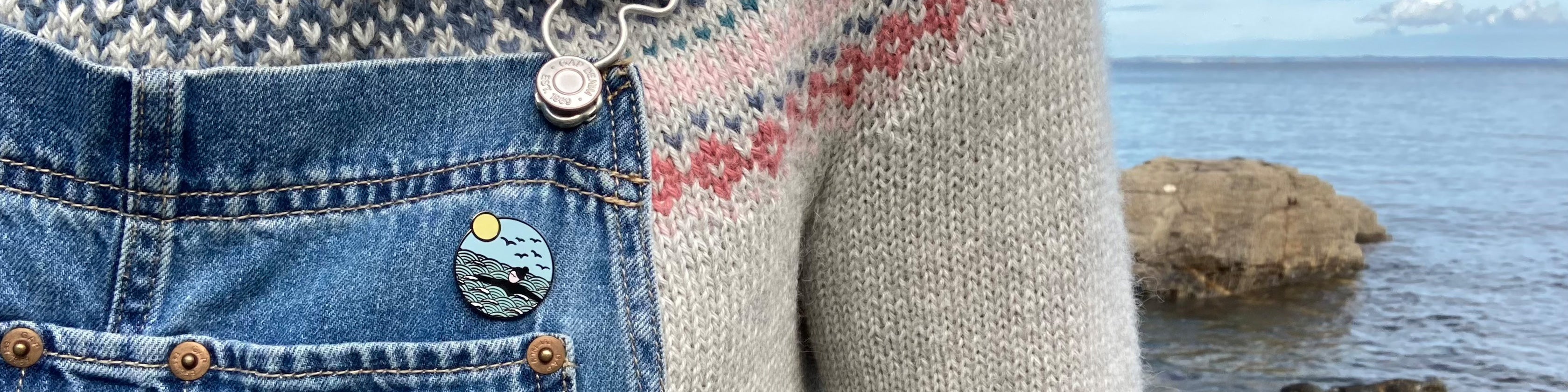 Close up of wild swimming enamel pin badge, attached to a pair of denim dungarees with a handknit fairisle jumper underneath. The sea can be seen behind the figure.