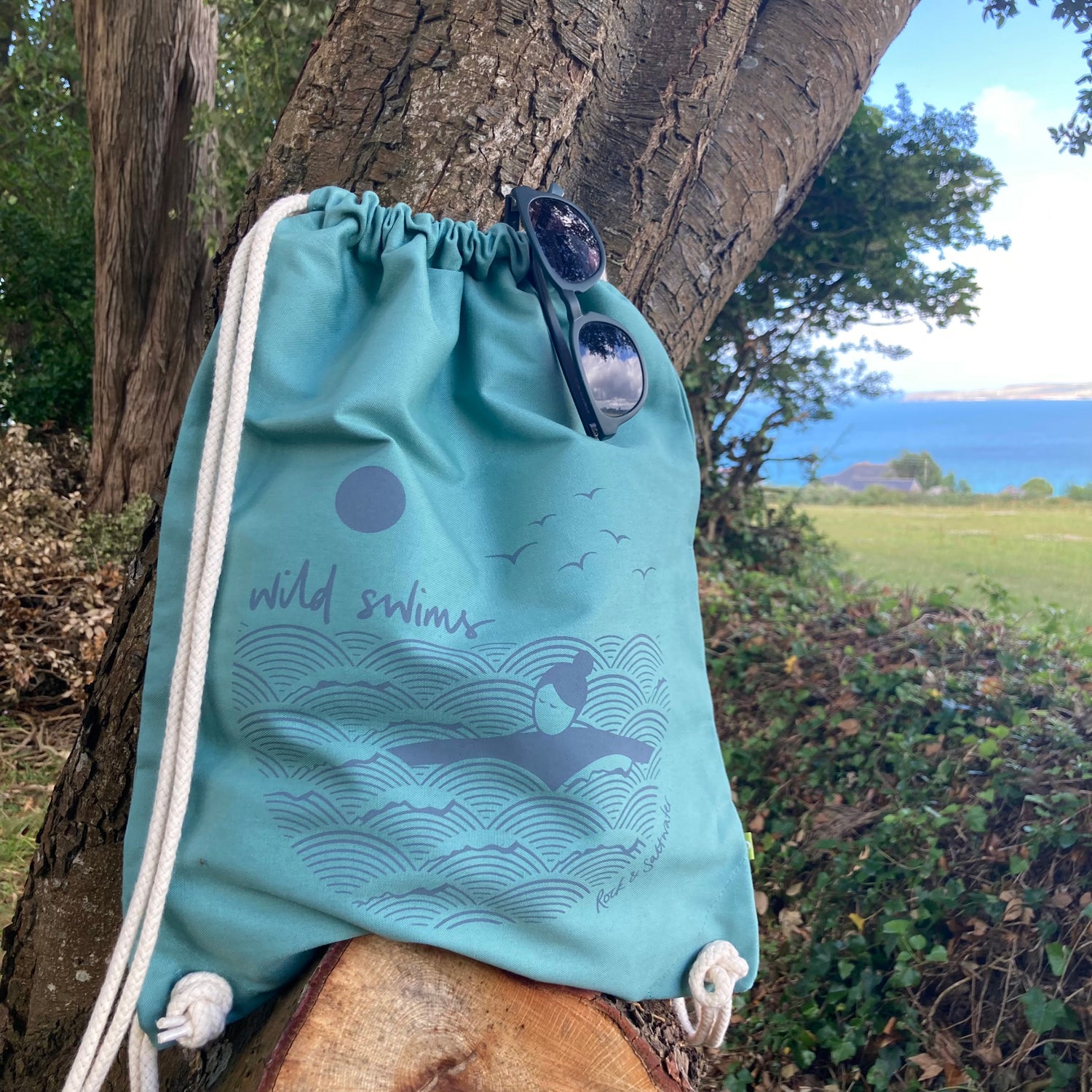 A sea green coloured drawstring gym bag rests against a tree with a view out towards the sea. The bag features a wild swimming illustration in dark grey, and a pair of sunglasses are on top of the bag.