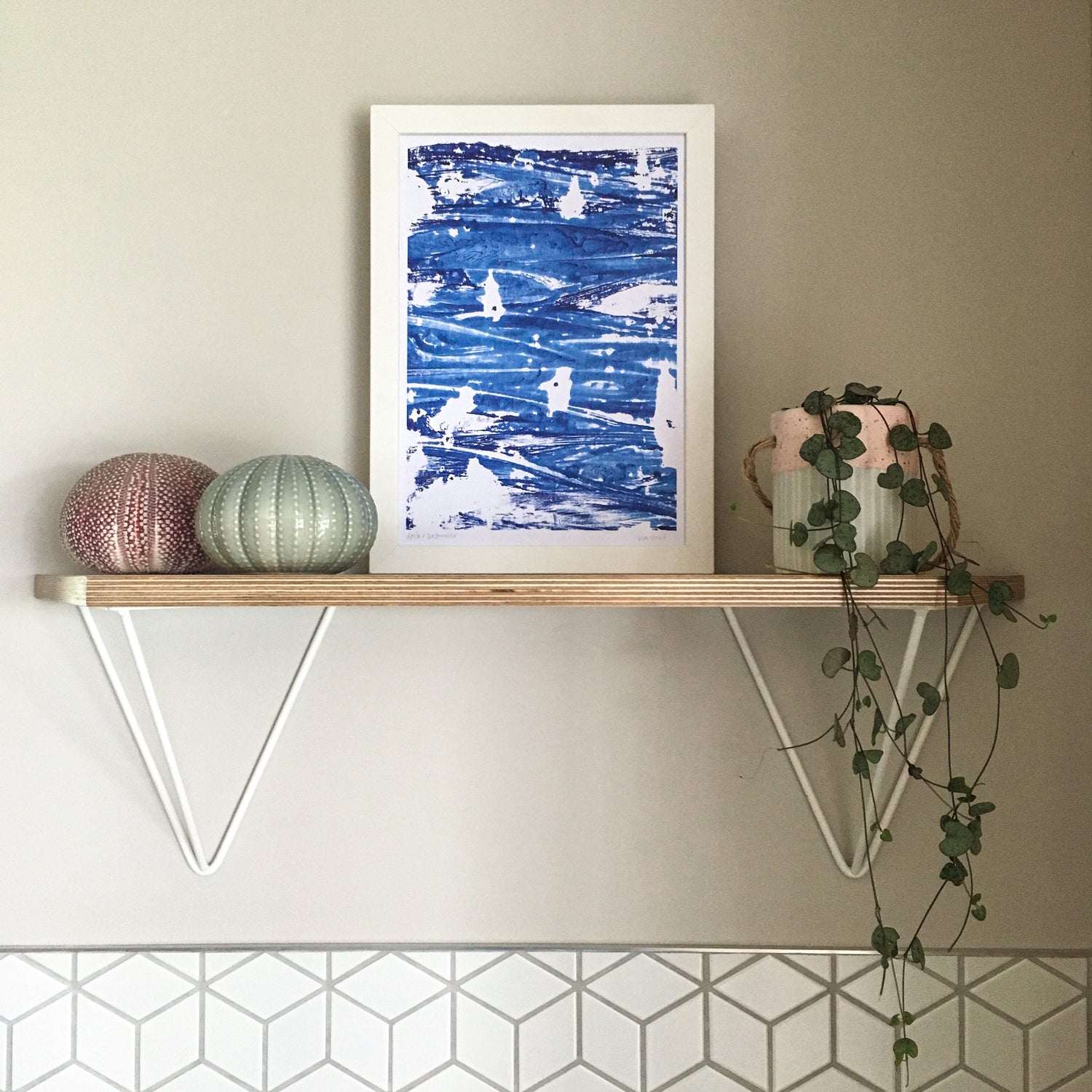 Wall with a birch ply and angled hairpin bracket shelf. On the shelf it a framed 'sea print' art print, two sea urchins (one ceramic) and a string of hearts plant in a ceramic pot. The wall is painted light grey with white geometric tiles below. 