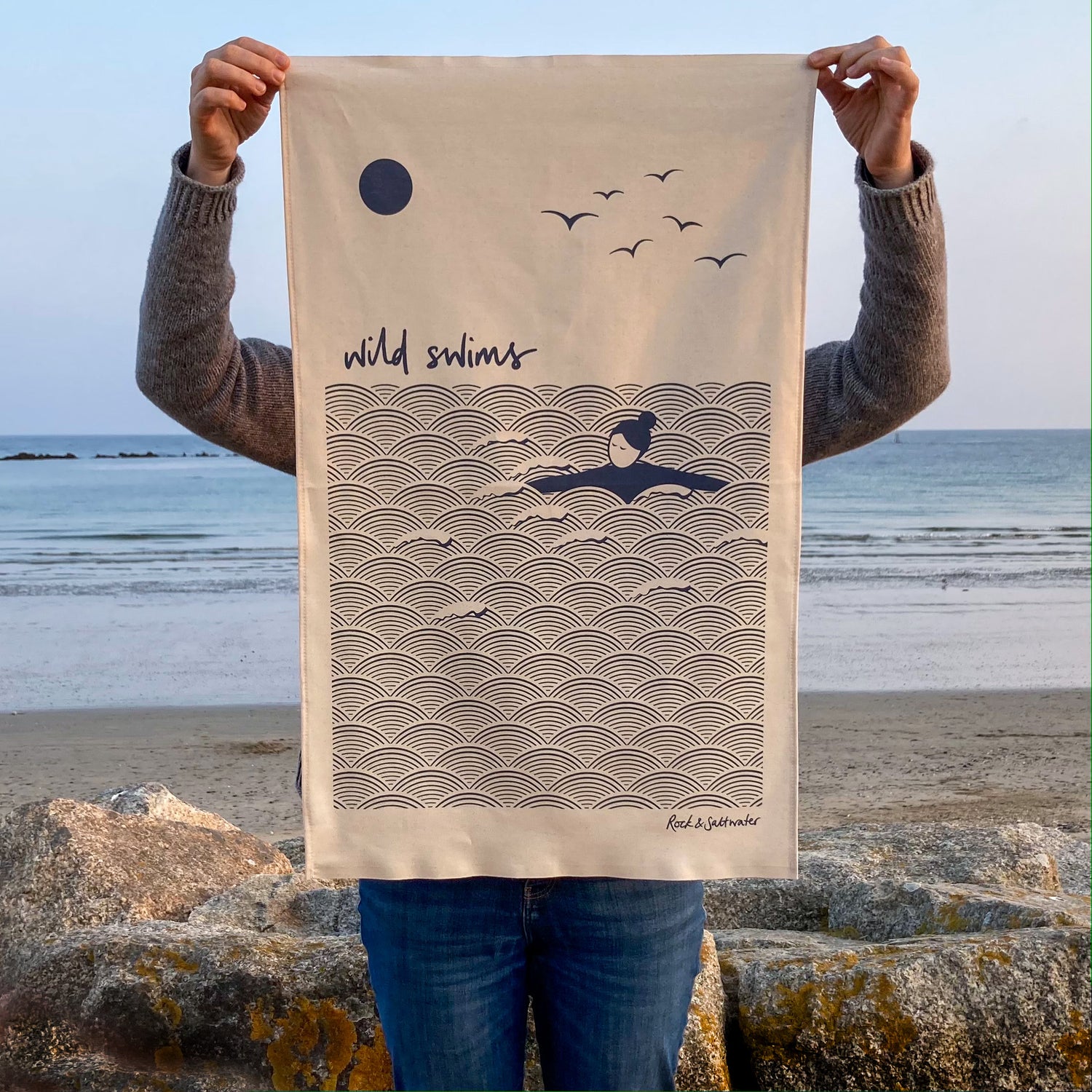 Women holding a 'wild swims' organic cotton tea towel, stood in front of the beach.