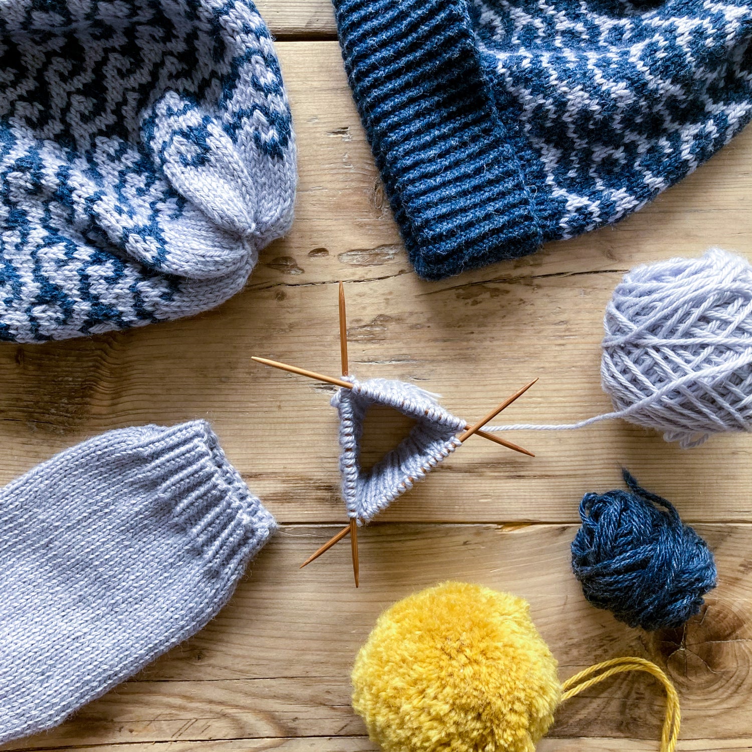 A rustic wooden table top with various knitted works in progress, including hats, pom poms and grey yarn on double pointed needles in the centre of the image.