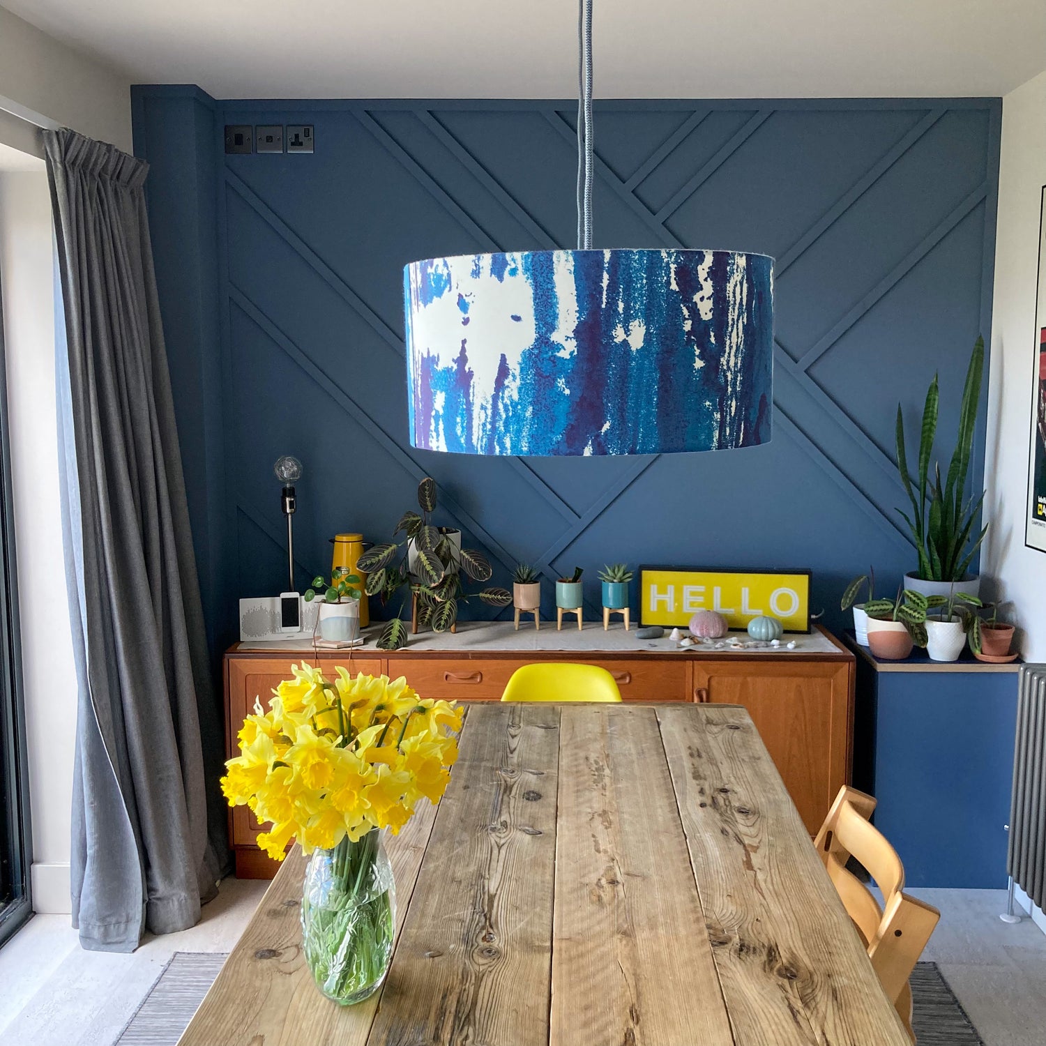 A dining table scene – a sea print hanging pendant lampshade hangs centrally over a scaffold board table with a vase full of daffodils sat on it. At the end of the room there is a dark blue feature wall, mid century sideboard and various house plants.