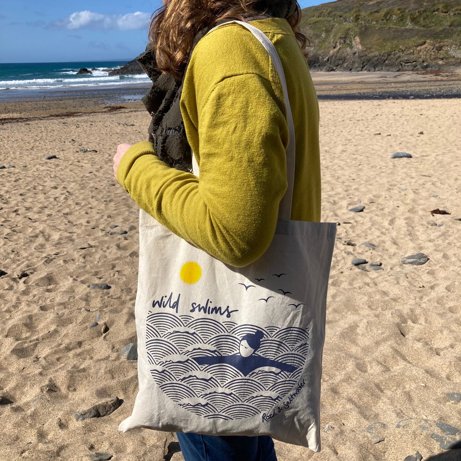 Figure stood on the beach looking out to see, wearing a yellow top with a natural 'wild swims' cotton tote bag over her shoulder. 