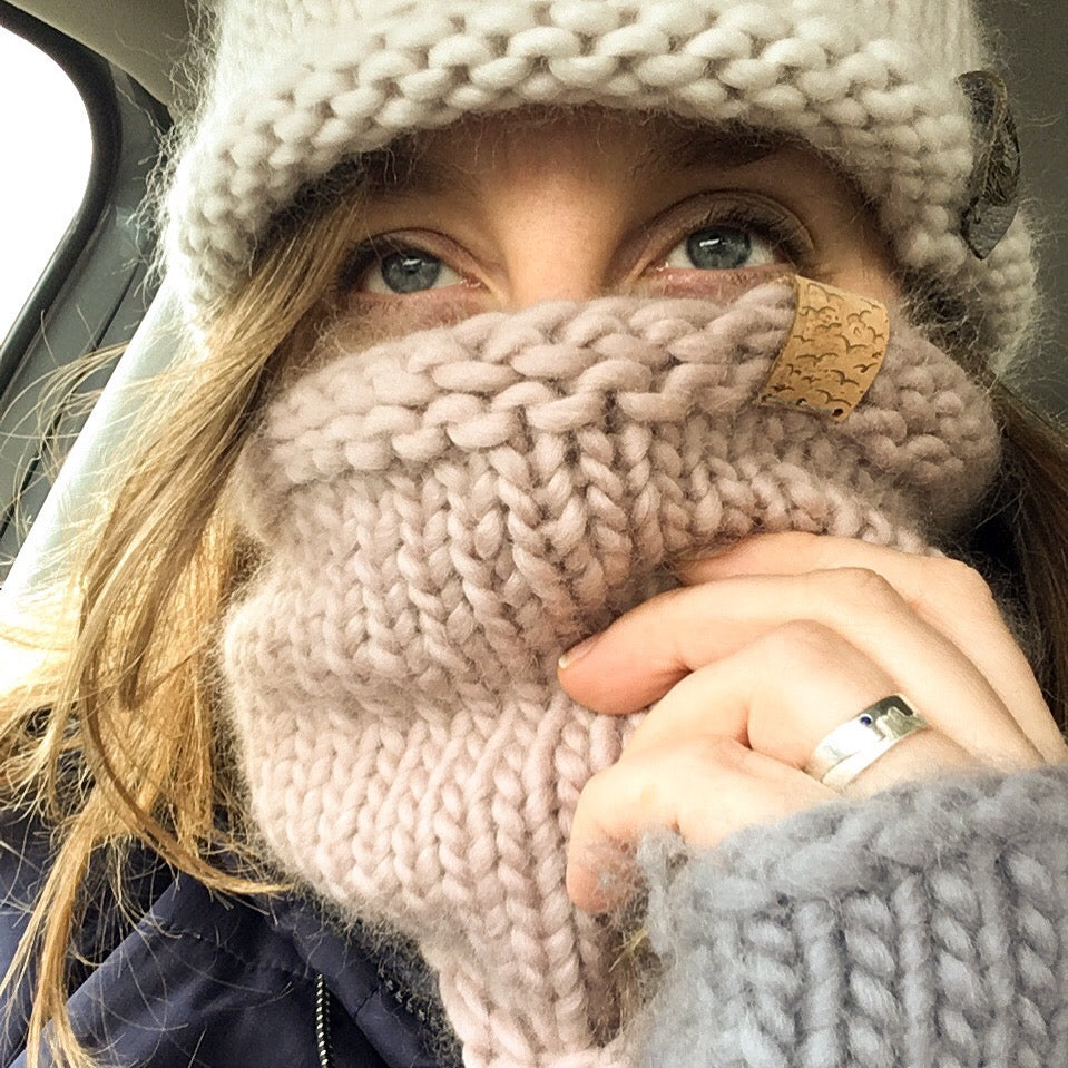 Woman wearing a hand knitted white hat, light pink cowl and light grey wrist warmer just visible. The knitted cowl is pulled up over her face so that just her eyes are visible.