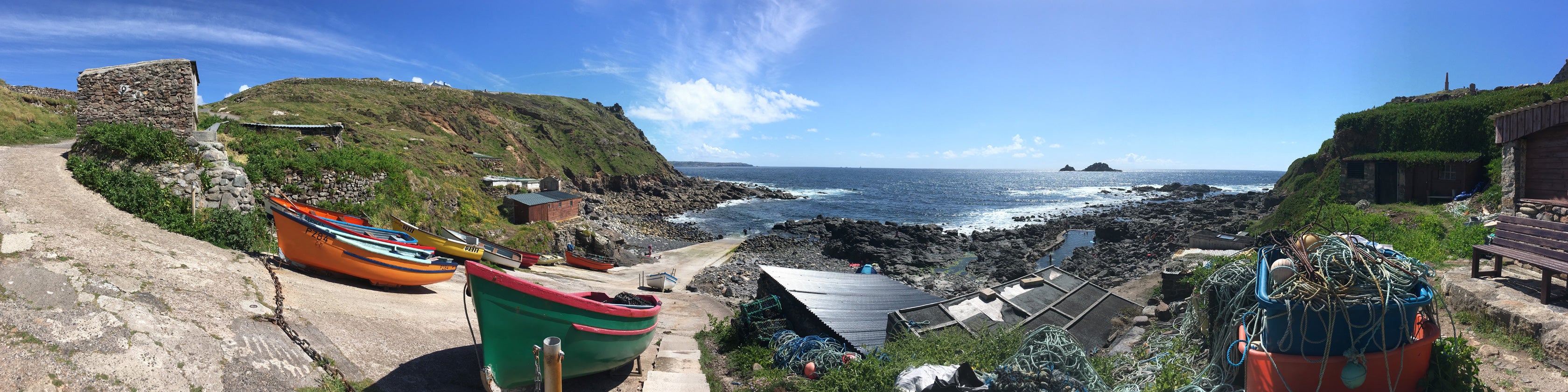 Panoramic photograph of a Cornish fishing cove, with fishing boats and nets in the foreground. A slipway leads down the the sea, the sky is blue with the odd cloud.